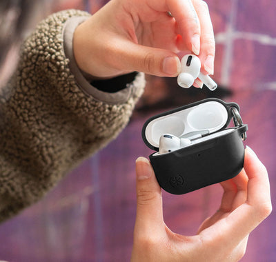 Woman putting an AirPods earbud into AirPods case with a Speck Presidio with Soft-Touch Coating case on it