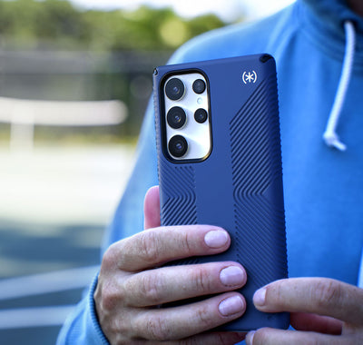 Man holding a Samsung Galaxy S23 Ultra in a Speck Presidio2 Grip case while standing on a tennis court